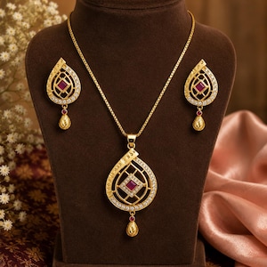 Puede incluir: Un conjunto de joyería en tono dorado que consta de un collar y pendientes a juego. Cada pieza presenta un marco en forma de lágrima con una piedra roja de corte cuadrado en el centro, rodeada de pequeños cristales blancos y un dije.