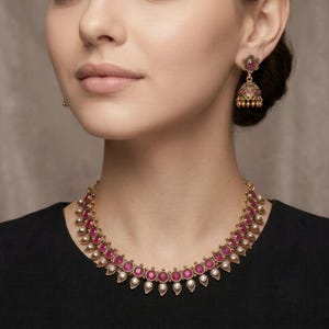 May include: A gold-toned necklace and matching jhumka earrings set featuring round, deep pink stones set in metal frames. The necklace has a repeating pattern of pink stones and small white accents, while the earrings feature a floral top.