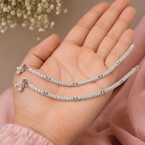 May include: A pair of silver-toned anklets featuring a textured chain design with small, colourful circular accents embedded along the length. Each anklet terminates with a small silver-toned clasp and a cluster of three tiny dangling bells.
