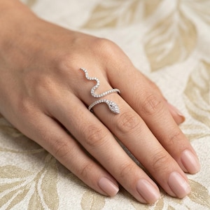 May include: A silver-toned snake ring featuring a winding body and head encrusted with small, clear, sparkling stones. The ring wraps around the finger, with the snake's head resting on the middle knuckle and the tail curving upwards.