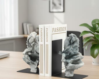 Könnte beinhalten: Ein Paar Buchstützen mit unregelmäßigen, strukturierten grauen steinartigen Skulpturen auf flachen schwarzen Metallsockeln, die zwei weiße Bücher stützen, auf deren Buchrücken und Vorderseiten das Wort FASHION in Schwarz gedruckt ist.