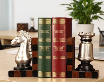 Könnte beinhalten: Ein Paar Buchstützen mit einer silberfarbenen Springer- und Turm-Schachfigur auf einem schwarz-braunen Schachbrettmuster-Sockel, die zwei gebundene Bücher mit grünen und roten Buchrücken und goldener Schrift stützen.