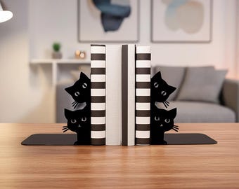 May include: Black metal cat-shaped bookends with four cat silhouettes peeking out from behind books. The bookends are on a wooden table, holding books with striped and solid covers. The background includes artwork and a couch.