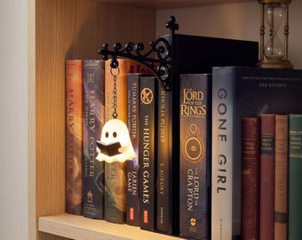 Könnte beinhalten: Ein schwarzer Buchständer aus Metall im Wandleuchten-Stil mit einer verzierten Halterung. Eine kleine, weiß leuchtende Geisterfigur, die ein winziges schwarzes Buch hält, hängt an einer dünnen Metallkette an der Halterung zwischen mehreren Büchern.