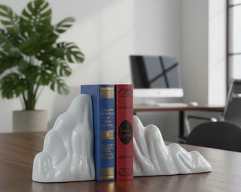 Könnte beinhalten: Ein Paar Buchstützen aus weißer Keramik mit einer glatten, wellenförmigen Bergform. Die Buchstützen sind auf beiden Seiten von zwei Hardcover-Büchern platziert, eines blau mit goldener Schrift und eines rot mit schwarzem und goldenem Text.