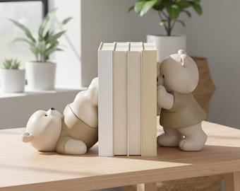 Könnte beinhalten: Ein Set aus zwei cremefarbenen Bären-Buchstützen in beigen Hemden. Ein Bär liegt auf dem Rücken und drückt seine Füße gegen einen Stapel aus drei Büchern, während der andere Bär aufrecht steht und gegen die gegenüberliegende Seite drückt.