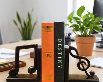 Könnte beinhalten: Ein Paar dunkelbraune Buchstützen aus Holz in Form von Musiknoten, die zwei Bücher aufrecht halten. Ein Buch hat einen leuchtend orangefarbenen Buchrücken mit Goldverzierungen, das andere ist ein schwarzes Buch mit dem Wort DESTINY in Weiß.