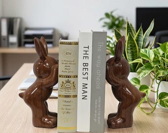 Könnte beinhalten: Ein Paar braune, glänzende Buchstützen in Hasenform mit Holzmaserung, die jeweils einen Hasen mit den Pfoten vor den Augen darstellen. Die Buchstützen stehen beidseitig von drei Büchern mit den Titeln "A Treasury of World's Classics", "THE BEST MAN" und "The way ahead".