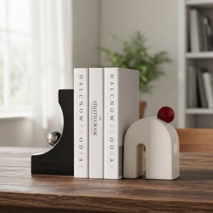 Puede incluir: Un juego de sujetalibros modernos en negro y crema. Un sujetalibros negro tiene un acento de bola plateada, y el otro es de color crema con una esfera roja encima. Tres libros con cubiertas blancas están entre ellos.