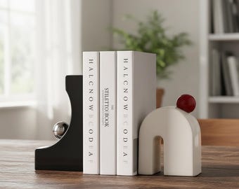 Könnte beinhalten: Ein Set moderner Buchstützen in Schwarz und Creme. Eine schwarze Buchstütze hat einen silbernen Kugelakzent, die andere ist cremefarben mit einer roten Kugel. Drei Bücher mit weißen Einbänden stehen dazwischen.