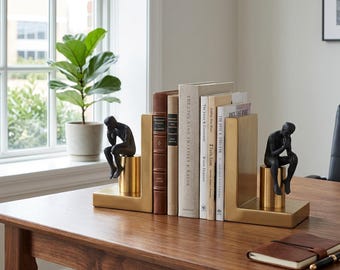 Könnte beinhalten: Ein Satz goldfarbener Metall-Buchstützen mit einer schwarzen Skulptur einer sitzenden, denkenden Person. Die Buchstützen halten eine Sammlung von Büchern mit verschiedenfarbigen Buchrücken. Eine Topfpflanze ist im Hintergrund zu sehen.