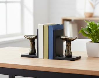 Könnte beinhalten: Ein Paar schwarze L-förmige Buchstützen mit bronzefarbenen Pilzskulpturen auf der Vorderseite. Die Buchstützen halten drei Bücher mit weißen, olivgrünen und blauen Einbänden, die vertikal zwischen den beiden Metallstützen angeordnet sind.