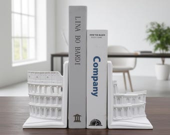 Könnte beinhalten: Weiße, kolosseumförmige Buchstützen halten zwei Bücher. Die Buchstützen stehen auf einer dunkelbraunen Oberfläche. Die Bücher tragen die Titel "Lina Bo Bardi" und "Company". Die Buchstützen sind ein dekorativer Artikel.