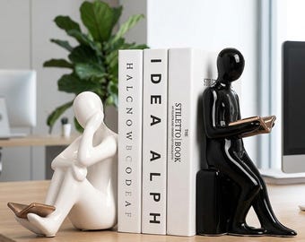 May include: A set of two ceramic bookends featuring stylized human figures in glossy white and black finishes. The white figure sits with a book on its feet, while the black figure sits on a block reading a book. Three white books are placed between them.