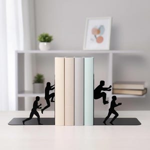 May include: Black metal bookends featuring silhouette figures of people climbing and jumping. The bookends support a row of books with beige, grey, and light blue covers. A decorative and functional item for a bookshelf.