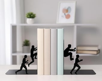 May include: Black metal bookends with silhouette figures of people climbing and jumping. The bookends hold a row of books with beige, gray, and light blue covers. A decorative and functional item for any bookshelf.