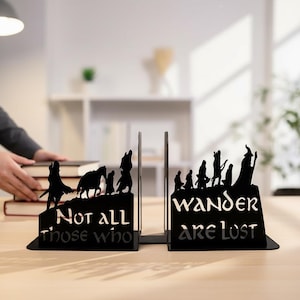 May include: Black metal bookends with a silhouette design featuring figures and the text "Not all those who" and "Wander are lost". The bookends are on a light-coloured surface.