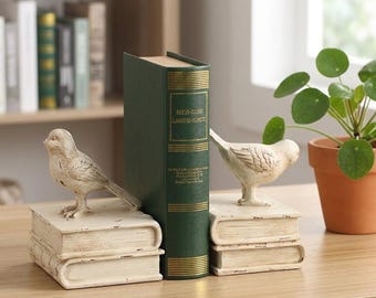 Könnte beinhalten: Ein Paar cremefarbene Buchstützen, die jeweils einen skulptierten Vogel auf einem Stapel von zwei Büchern zeigen. Die Buchstützen haben ein abgenutztes Finish und stehen auf beiden Seiten eines grünen Buches mit goldener Schrift am Buchrücken.