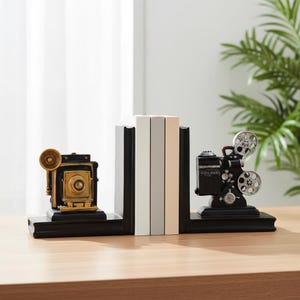 May include: A pair of black bookends featuring a vintage gold-toned bellows camera on the left and a black and silver film projector on the right. The bookends support three books with white, light gray, and beige spines, resting on a flat surface.