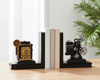 Könnte beinhalten: Ein Paar schwarze Buchstützen mit einer goldfarbenen Vintage-Faltenbalgkamera auf der linken Seite und einem schwarz-silbernen Filmprojektor auf der rechten Seite. Die Buchstützen halten drei Bücher mit weißen, hellgrauen und beigen Buchrücken.