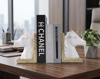 Könnte beinhalten: Weiße Einhorn-Buchstützen mit goldenen Hörnern und Sockeln flankieren einen Stapel Bücher. Die Buchstützen stehen auf einer dunklen Holzoberfläche. Die Bücher tragen die Aufschriften "CHANEL", "FULL STOP" und "SENIOR ASH". Eine kleine Topfpflanze ist sichtbar.