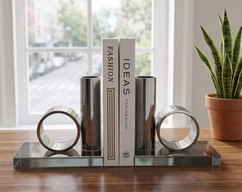 Könnte beinhalten: Ein modernes Buchstützen-Set mit zwei silbernen zylindrischen Stützen und zwei silbernen Ringakzenten, die auf einer rechteckigen Glasbasis ruhen. Zwei Bücher mit den Titeln "FASHION" und "IDEAS TOP DESIGN" werden zwischen den Stützen gehalten. Eine Topfpflanze befindet sich im Hintergrund.