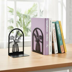 Puede incluir: Un par de sujetalibros de metal negro con un diseño recortado de un jarrón lleno de flores. Un sujetalibros está solo, mientras que el otro sostiene una fila de cinco libros con lomos de varios colores, incluyendo morado, blanco, turquesa y amarillo.