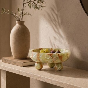 Peut inclure: Un bol &agrave; fruits d&eacute;coratif en onyx vert et marron, avec trois pieds arrondis. Il contient des raisins verts et des figues. Un vase beige textur&eacute; et un livre intitul&eacute; "Picasso" sont &eacute;galement visibles.