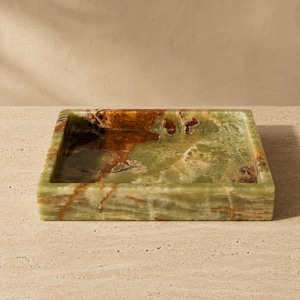 May include: A square tray crafted from polished green onyx stone, featuring natural veining in shades of deep brown, cream, and translucent light green. The tray has a smooth, flat base and raised edges, showcasing the unique mineral patterns.