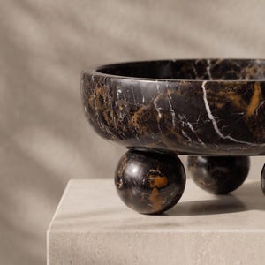 May include: A circular bowl crafted from dark brown and black marble with prominent white veining. The bowl features a smooth, polished finish and is supported by three spherical marble feet that elevate the main basin above the surface.