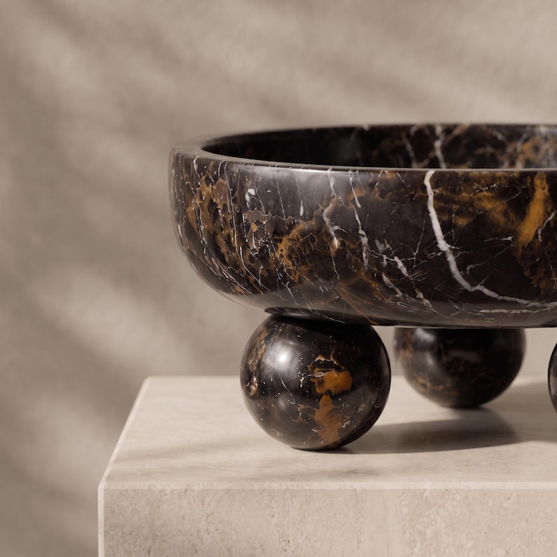 May include: A circular bowl crafted from dark brown and black marble with prominent white veining. The bowl features a smooth, polished finish and is supported by three spherical marble feet that elevate the main basin above the surface.