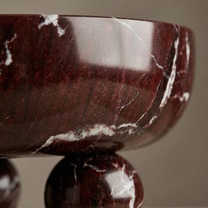 Pu&ograve; includere: Una ciotola decorativa in marmo rosso con venature bianche. La ciotola &egrave; sostenuta da tre piedi sferici. La superficie lucida riflette la luce, evidenziando i motivi e i colori naturali della pietra.
