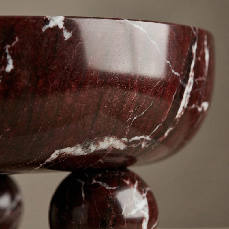 Pu&ograve; includere: Una ciotola decorativa in marmo rosso con venature bianche. La ciotola &egrave; sostenuta da tre piedi sferici. La superficie lucida riflette la luce, evidenziando i motivi e i colori naturali della pietra.