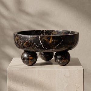 May include: A circular bowl crafted from black marble with prominent white and gold veining. The bowl features a smooth, polished finish and is supported by three spherical feet, also made of matching black marble with similar veining patterns.