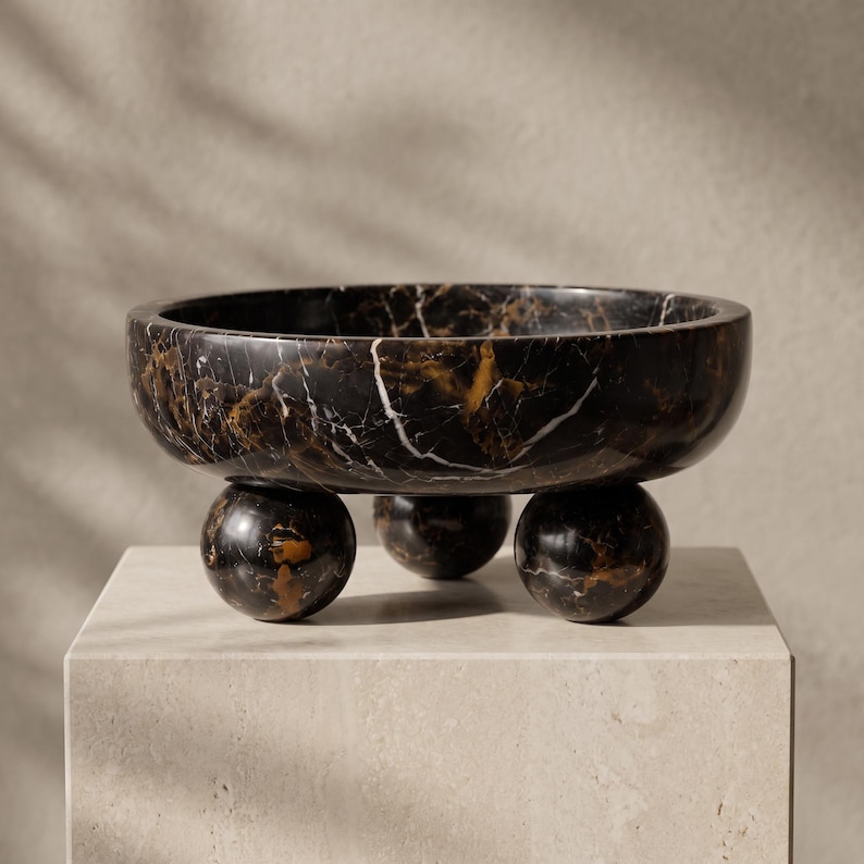 May include: A circular bowl crafted from black marble with prominent white and gold veining. The bowl features a smooth, polished finish and is supported by three spherical feet, also made of matching black marble with similar veining patterns.