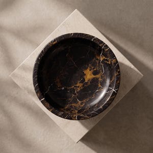 May include: A circular bowl crafted from dark black marble featuring prominent, intricate veining in shades of gold, amber, and white. The bowl has a smooth, polished finish and a deep, rounded interior basin, showcasing the natural stone pattern.