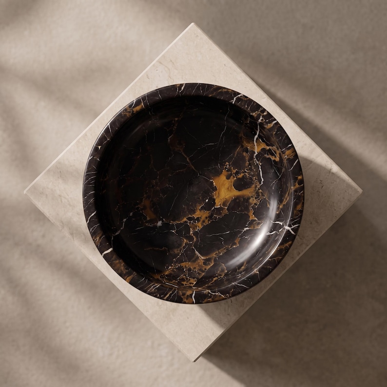 May include: A circular bowl crafted from dark black marble featuring prominent, intricate veining in shades of gold, amber, and white. The bowl has a smooth, polished finish and a deep, rounded interior basin, showcasing the natural stone pattern.
