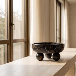 May include: A circular black marble bowl featuring prominent white and gold veining throughout the stone. The bowl is supported by three spherical marble feet, each matching the dark, polished stone texture of the main basin structure.