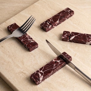Può includere: Quattro poggia posate rettangolari in marmo rosso scuro con venature bianche. Una forchetta d'argento poggia su un pezzo e un coltello d'argento su un altro. Due ulteriori pezzi di marmo sono posizionati vicino su una superficie beige.