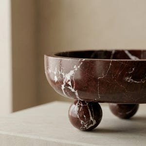 Pu&ograve; includere: Una ciotola in marmo bordeaux con venature bianche, appoggiata su tre piedini sferici. La ciotola ha una forma arrotondata e una superficie liscia e lucida. Questo pezzo decorativo &egrave; ideale per esporre frutta o come centrotavola.