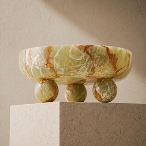 Peut inclure: Un bol d&eacute;coratif en onyx vert avec des veines marron. Le bol est soutenu par trois pieds sph&eacute;riques. Le bol est pr&eacute;sent&eacute; sur un pi&eacute;destal carr&eacute; de couleur claire. L'onyx a une qualit&eacute; translucide.