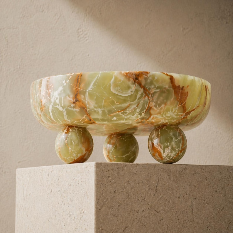 Peut inclure: Un bol d&eacute;coratif en onyx vert avec des veines marron. Le bol est soutenu par trois pieds sph&eacute;riques. Le bol est pr&eacute;sent&eacute; sur un pi&eacute;destal carr&eacute; de couleur claire. L'onyx a une qualit&eacute; translucide.