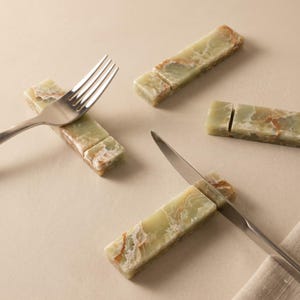 Op de afbeelding: Een verzameling groene en beige onyx messenleggers en een vork en mes. De rechthoekige leggers hebben een natuurstenen uitstraling met kleurvariaties. De roestvrijstalen vork en mes zijn op de leggers geplaatst.