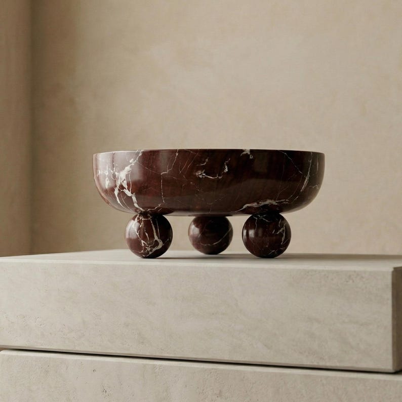 Pu&ograve; includere: Una ciotola in marmo bordeaux con venature bianche, appoggiata su tre piedi sferici. La ciotola ha un design ampio e poco profondo, perfetto per esporre oggetti decorativi o servire cibo. La superficie lucida riflette la luce.