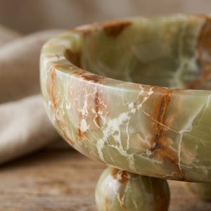 Peut inclure: Un bol en onyx vert avec des veines marron et blanches. Le bol a une forme arrondie et repose sur une base sph&eacute;rique. La surface polie refl&egrave;te la lumi&egrave;re, soulignant les motifs naturels de la pierre. Une pi&egrave;ce d&eacute;corative pour la maison.