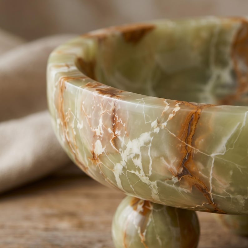 Peut inclure: Un bol en onyx vert avec des veines marron et blanches. Le bol a une forme arrondie et repose sur une base sph&eacute;rique. La surface polie refl&egrave;te la lumi&egrave;re, soulignant les motifs naturels de la pierre. Une pi&egrave;ce d&eacute;corative pour la maison.