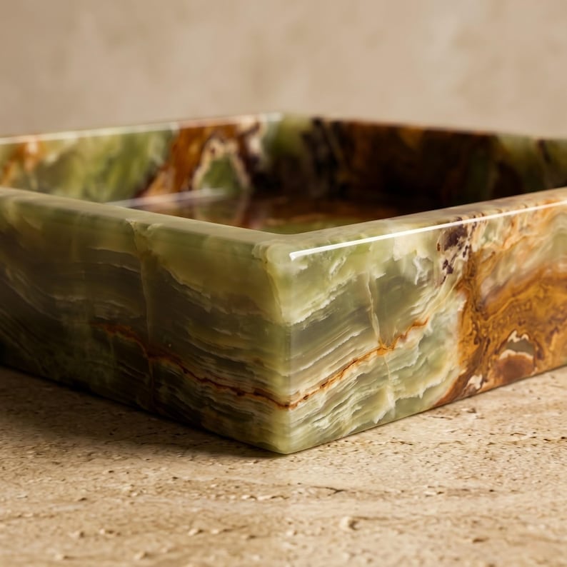 May include: A square bowl crafted from polished green onyx stone, featuring natural banded patterns in shades of sage green, cream, and deep amber. The stone surface displays a smooth, glossy finish with visible mineral veining throughout the structure.