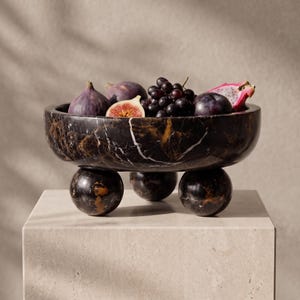May include: A black marble bowl with white and gold veining, supported by three spherical marble feet. The bowl is filled with fresh fruit, including whole purple figs, a sliced fig, a cluster of dark grapes, and a piece of pink dragon fruit.