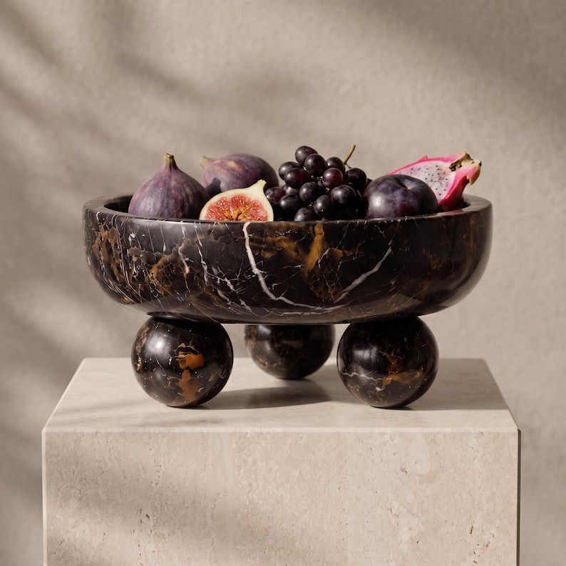May include: A black marble bowl with white and gold veining, supported by three spherical marble feet. The bowl is filled with fresh fruit, including whole purple figs, a sliced fig, a cluster of dark grapes, and a piece of pink dragon fruit.