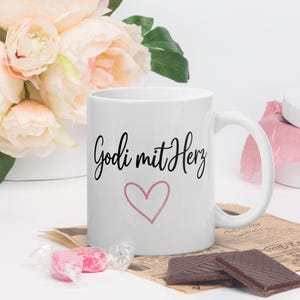 Könnte beinhalten: Weiße Keramik-Tasse mit dem Text "Godi mit Herz" in schwarzer Schrift und einem rosa Herz. Die Tasse steht auf einem Vintage-Papier mit Schokoriegeln und verpackten Süßigkeiten. Im Hintergrund ein Strauß rosa und weißer Blumen.